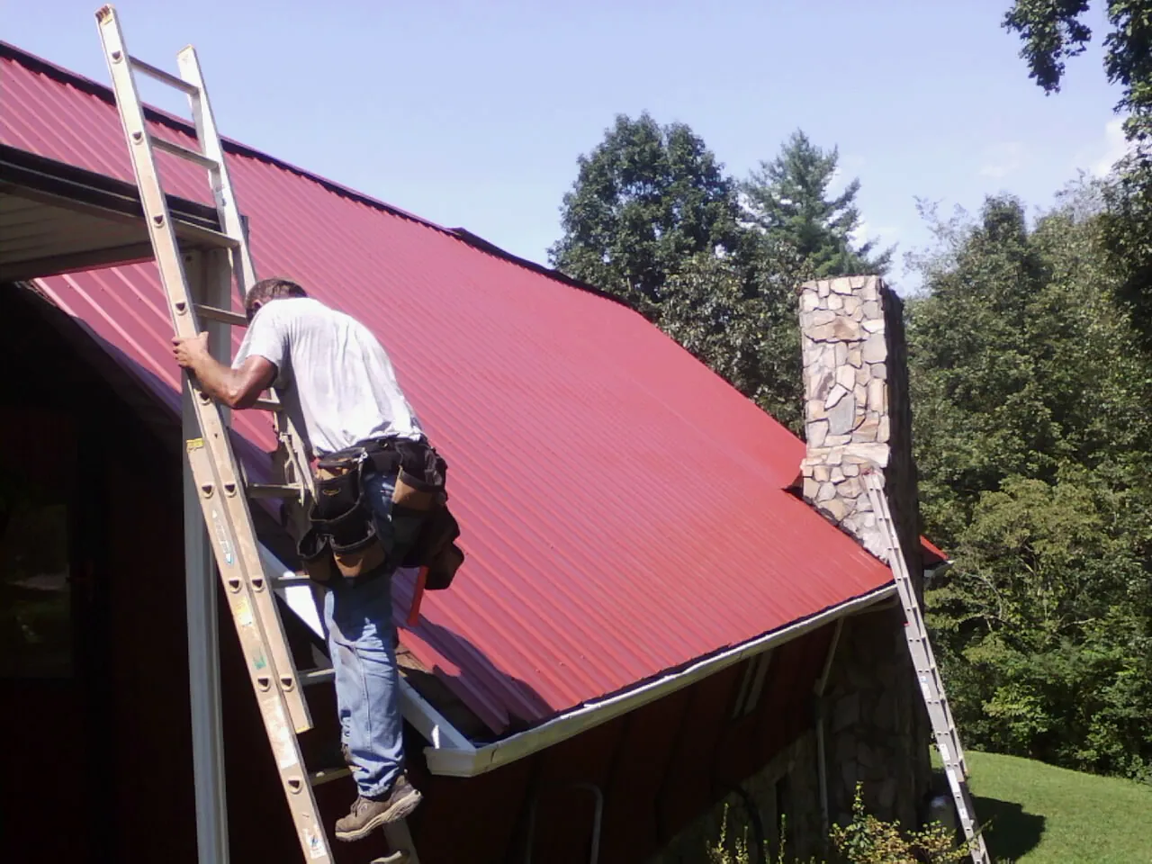 Roofer on a ladder beside a red metal roof and stone chimney — Soffit & Fascia Repair by Roofing Brothers in Littlestown